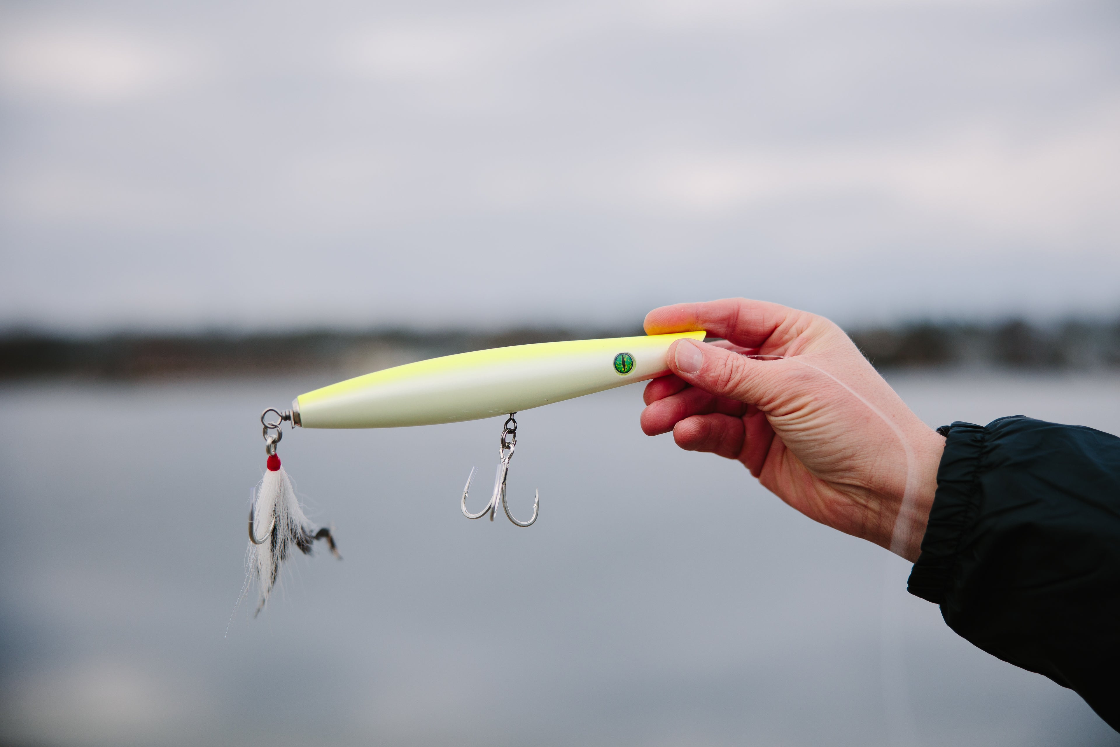 Hand holding a yellow fishing pencil popper lure with a blurred lake background