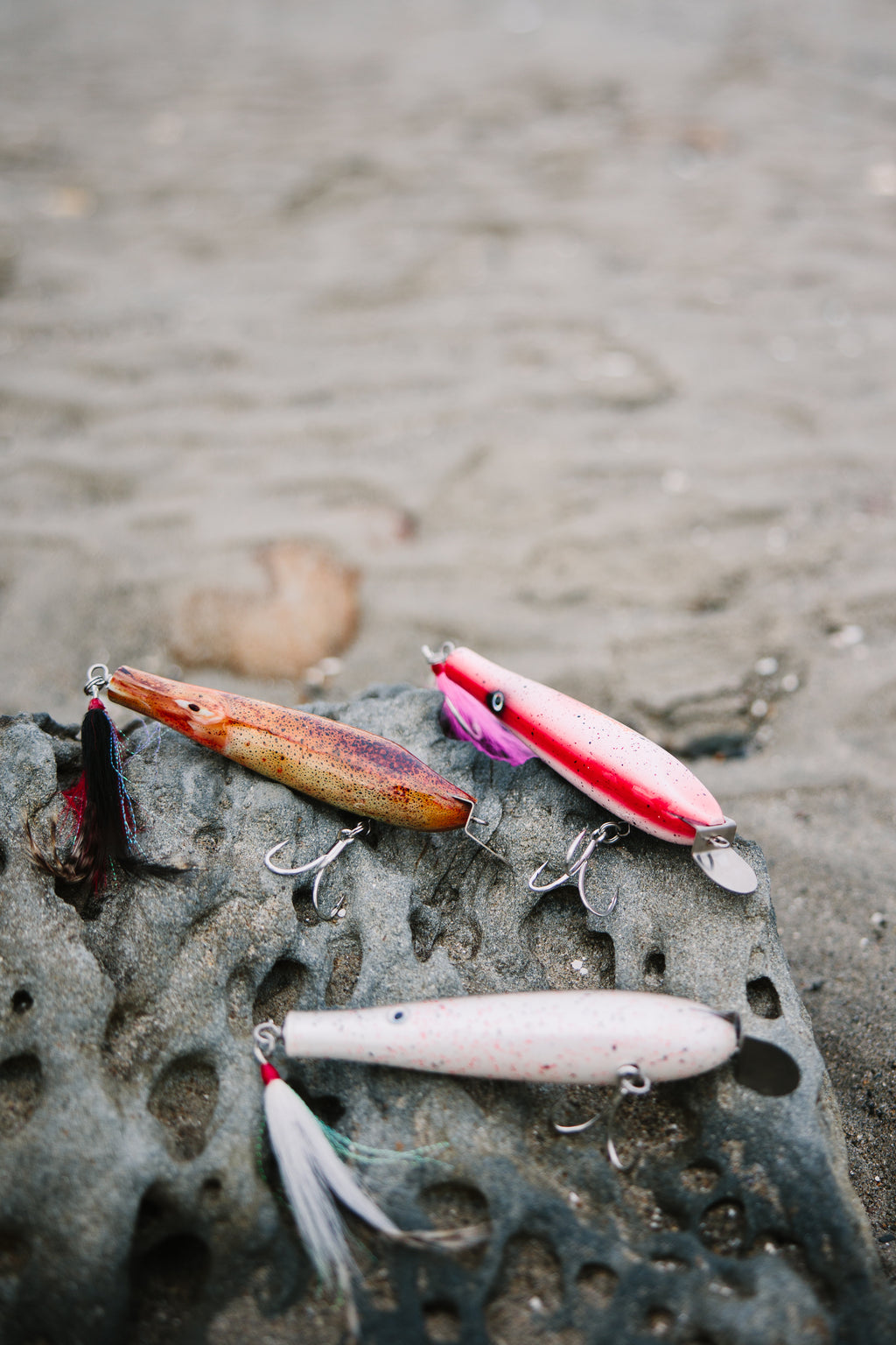 Three metal lip fishing lures on a rocky surface with a blurred sand background