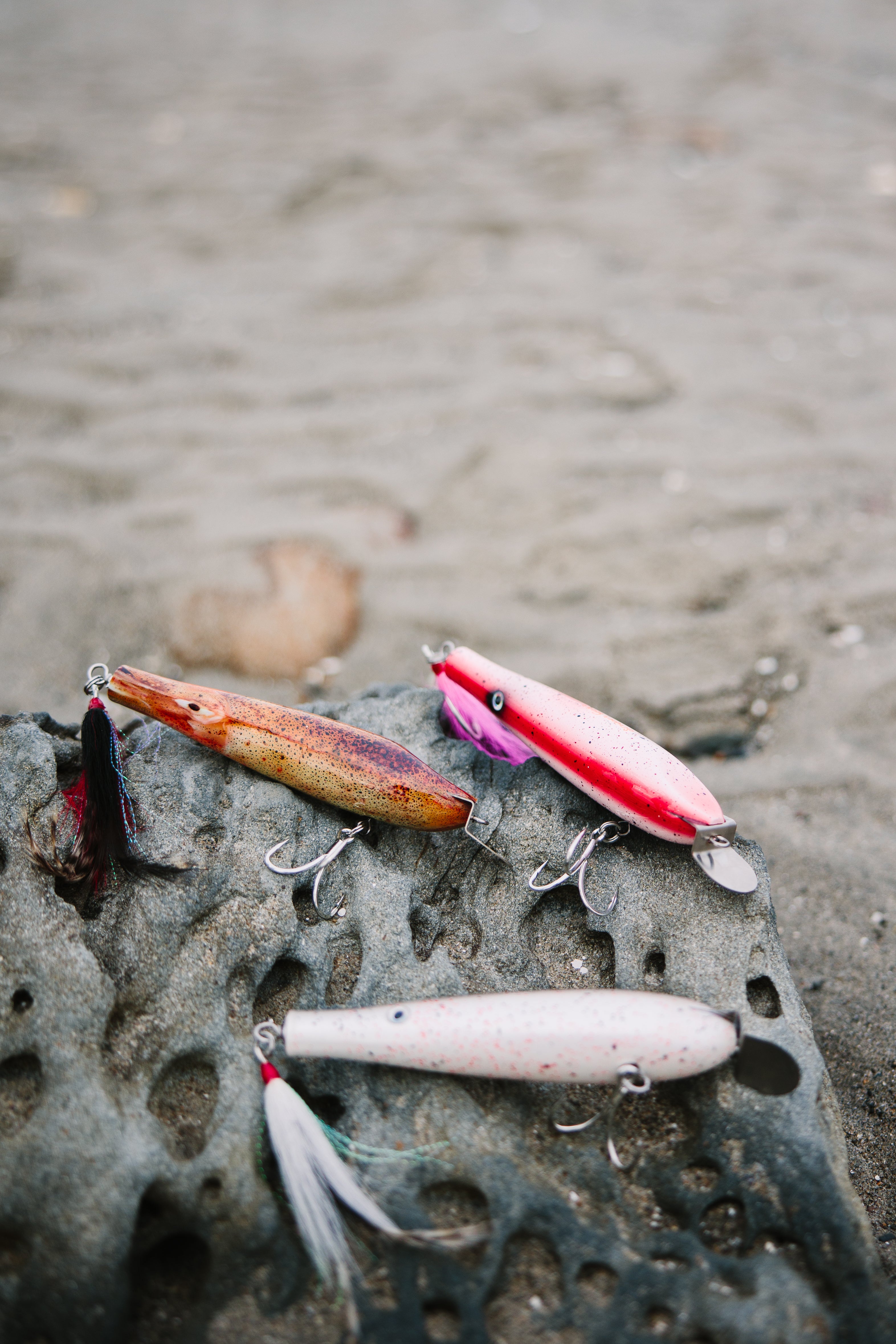 Three metal lip fishing lures on a rocky surface with a blurred sand background