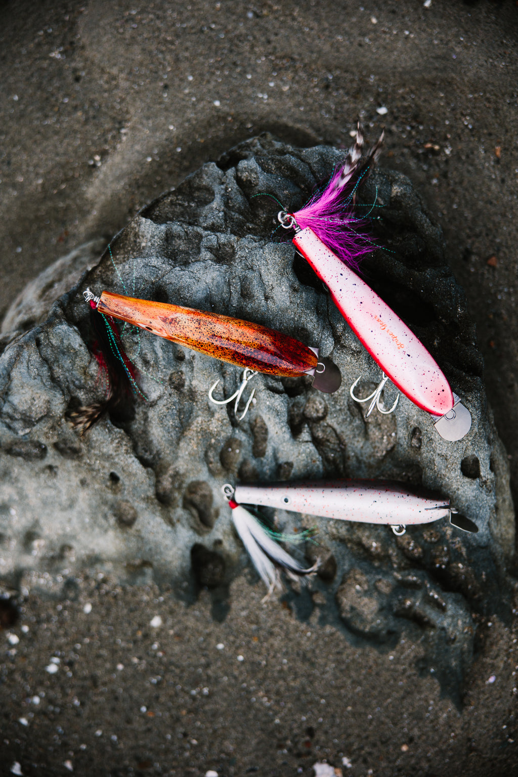 Three metal lip fishing lures on a rocky surface in the sand